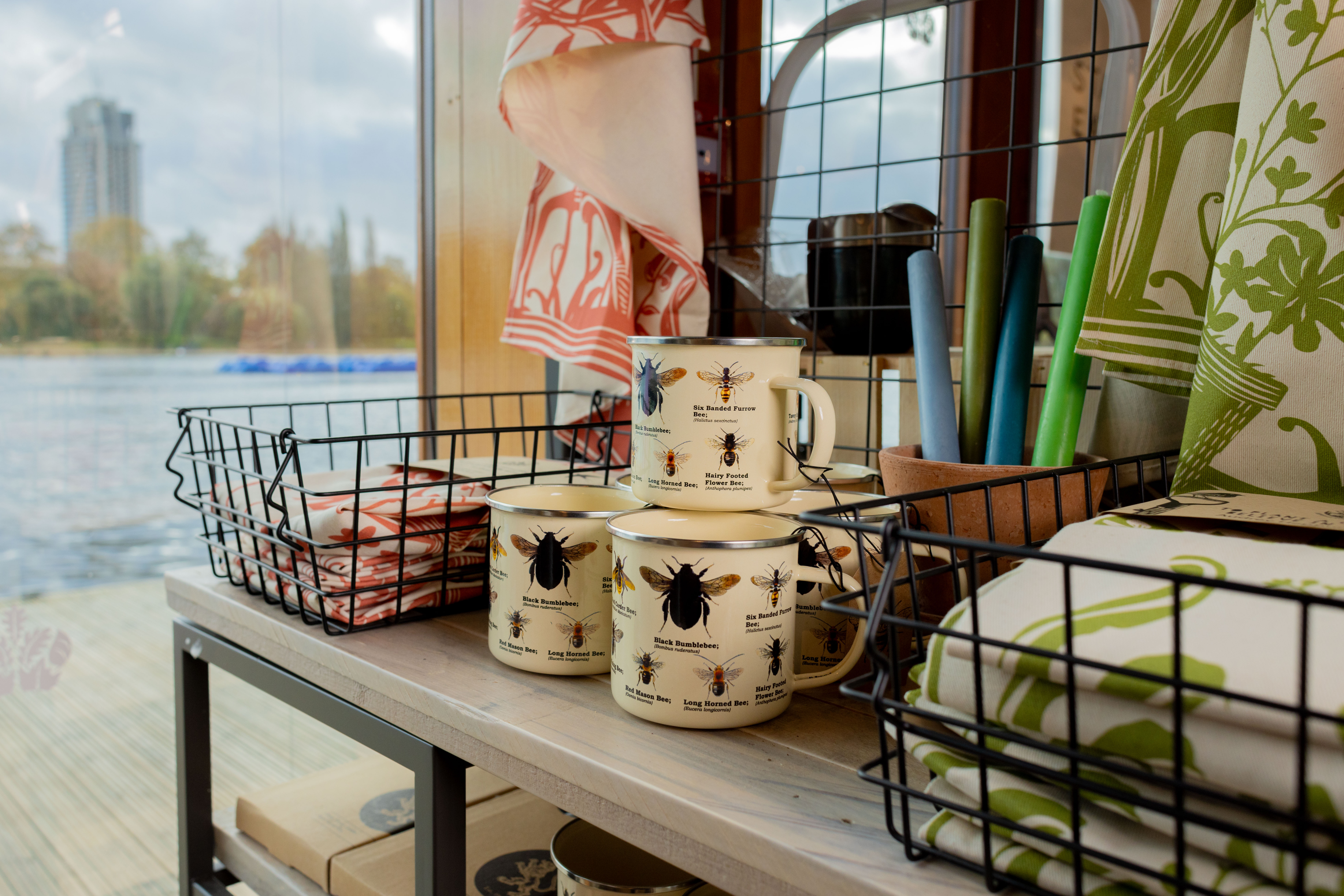 A shop display with the Serpentine lake in the background. A collection on mugs with bee illustrations on sits in the foreground. 