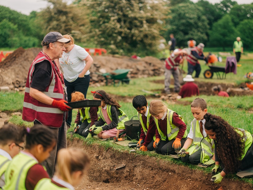 A volunteer taching a group of schoolchildren how to dig and look for find in a shallow trench. The volunteer is standing whilst the children are kneeling over the trench.