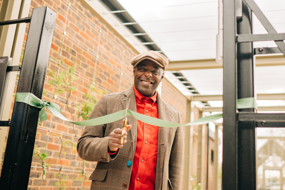 Paterson Joseph is seen looking at the camera whilst cutting a green ribbon across the cafe gate with some gold scissors