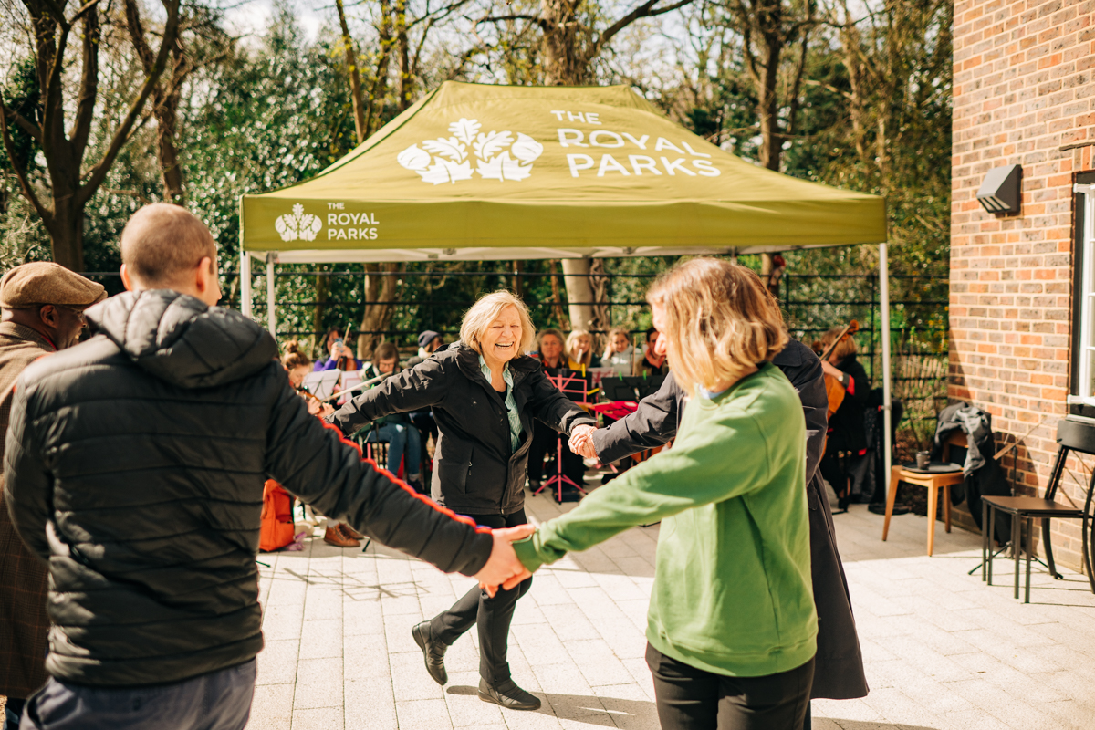 People are seen dancing in a ring outside the cafe with a Royal Parks branded gazebo in the background.