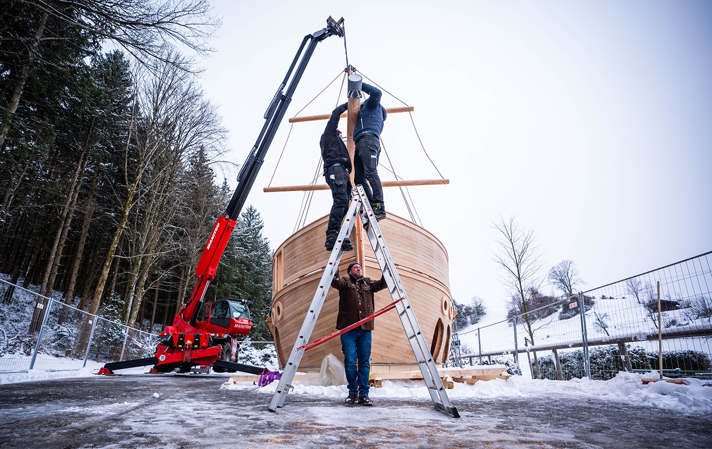 Two men work up a ladder to fit the bowsprit to the galleon, while a third man holds the ladder still. The ground and surrounding trees are snow-covered, and a crane holds the bowsprit in mid-air.