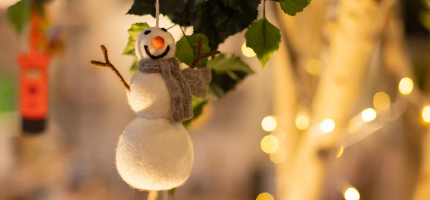 A snowman tree decoration made from felt hangs with fairy lights in the background