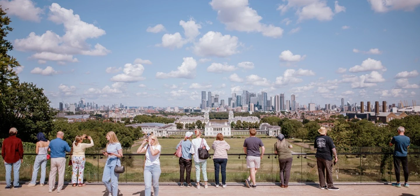 A view from the General Wofle statue looking down toward the Queen's House. White clouds pepper a blue sky and people stand with their back to the camera as they look down at the view