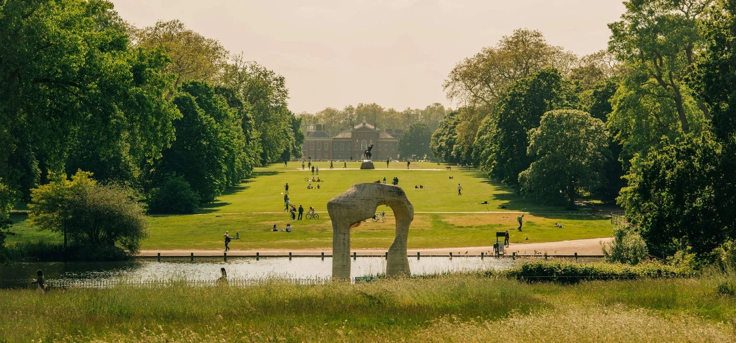 a wide shot of a green space with people and a lake in Kensington Gardens