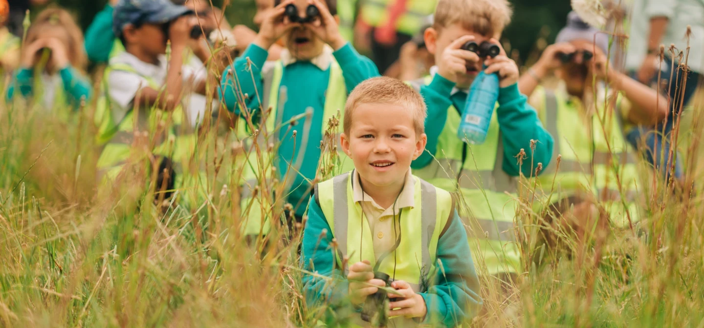 group of young children wearing high-visibility vests and school uniforms are exploring nature in a grassy field. Some of them are using binoculars to observe their surroundings, while one boy in the foreground smiles at the camera, holding binoculars in his hands. The background shows more children and adults, also dressed in high-visibility vests, engaged in the outdoor activity.