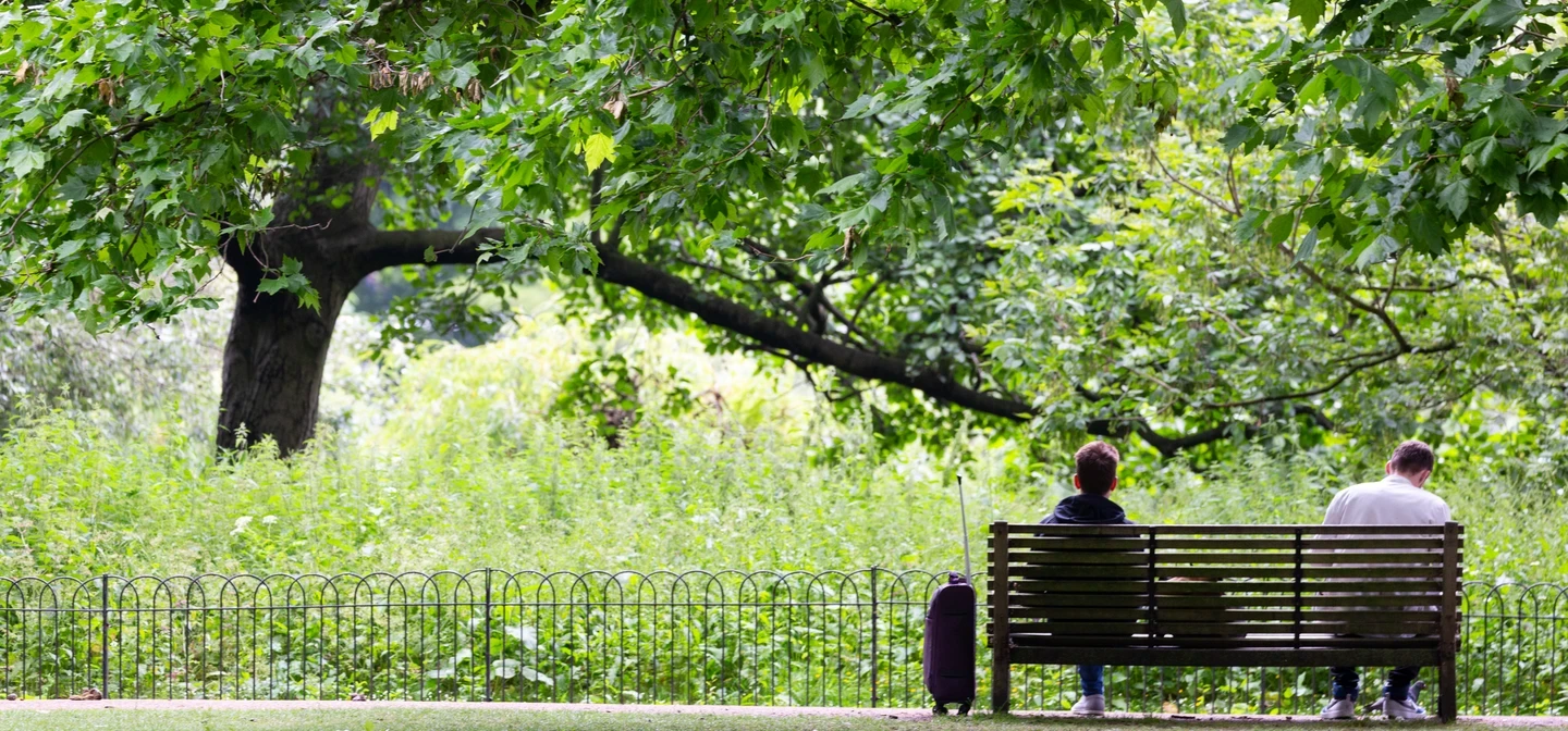 Image of two people sitting on a wooden bench, in a large, green outdoor space