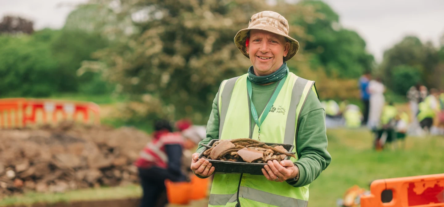 Image of an archaeologist holding onto historical artefacts in an outdoor, park setting with people working and digging in the background.