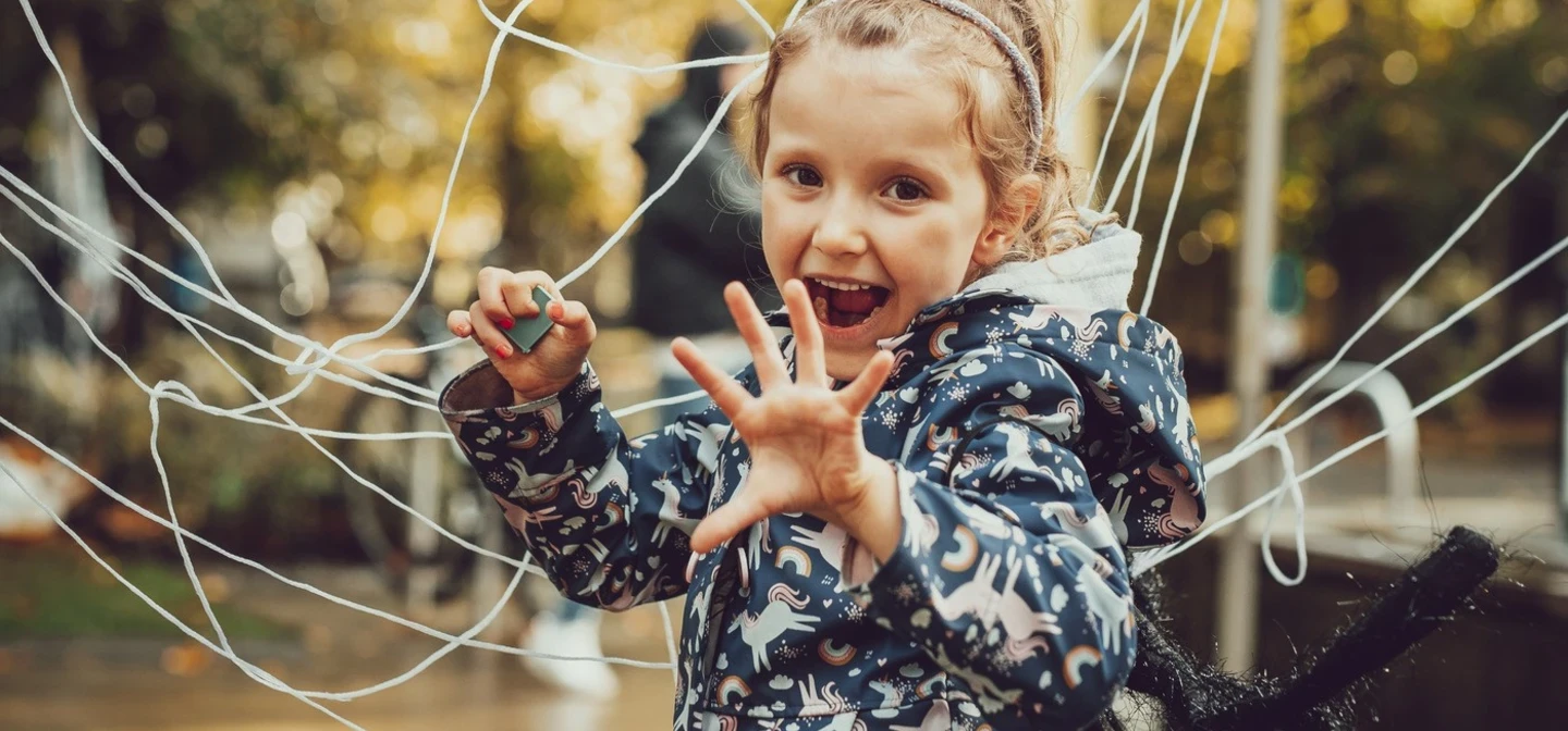 A girl, wearing a spider costume is doing a playful scary pose in front of a big cobweb made of string