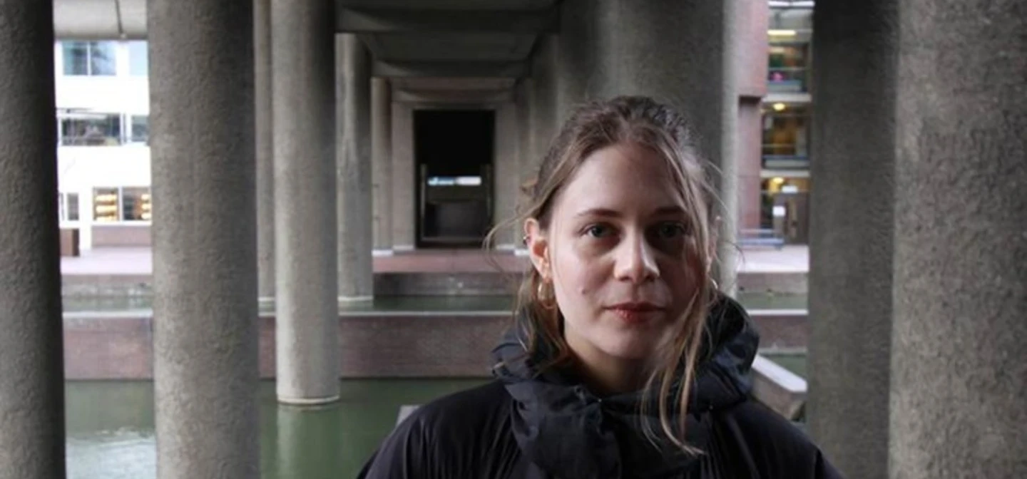 Leah, a white woman in her 30s, smiles and looks at the camera. She is wearing a large black coat and stands underneath a bridge or other structure supported by concrete pillars.