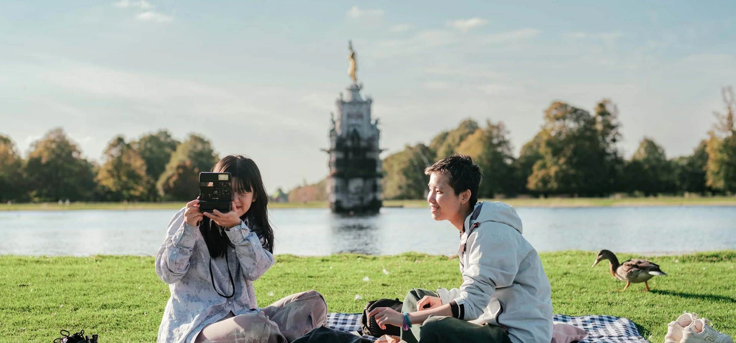 A couple picnicking and taking photography in Bushy Park, with the Diana fountain in the background