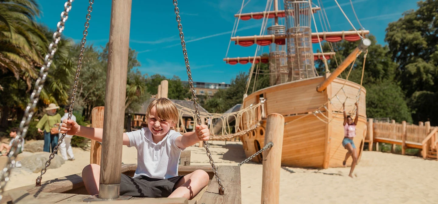 A boy sits in the wooden play boat with the new galleon behind