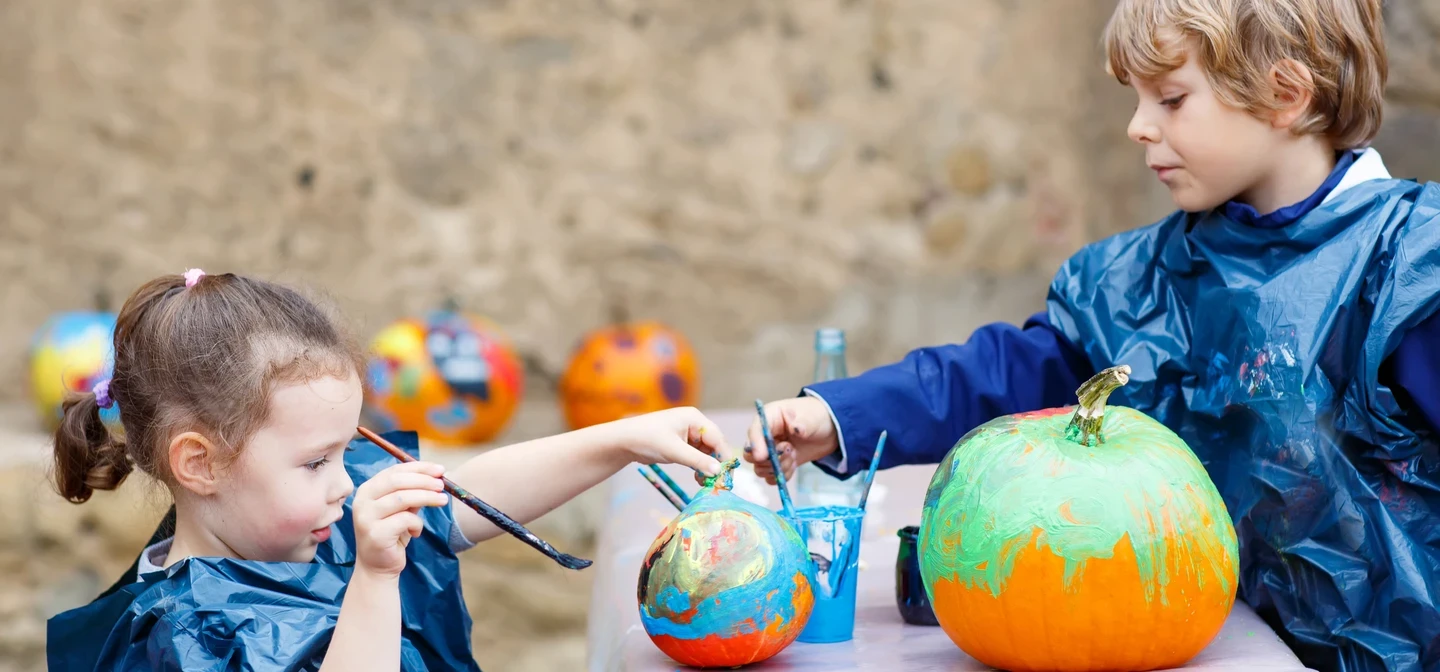 Two children sit either side of a table. They are wearing blue plastic aprons and have paintbrushes in their hands directed at large orange pumpkins that are being colourfully painted. 