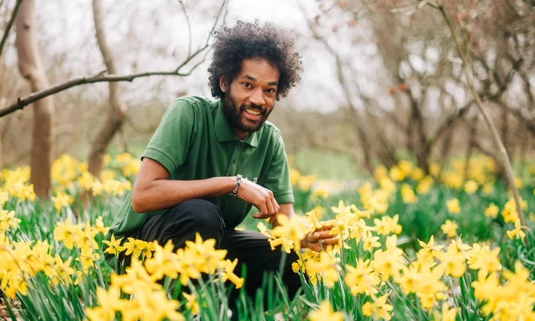 A young man smiling at the camera, bending down in a field of yellow daffodils.