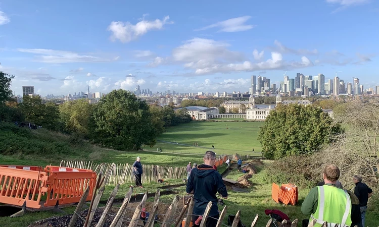 A view looking down the Grand Ascent over Greenwich Park toward Canary Wharf, it is a sunny day and the sky is blue as the volunteers start digging the trench in the foreground.