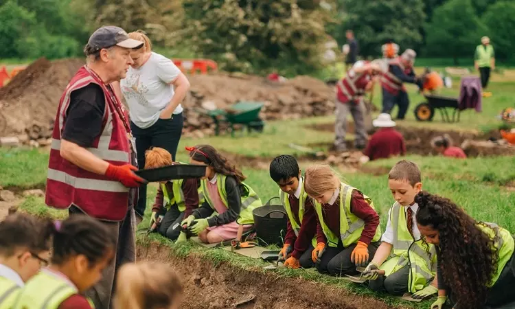 A volunteer taching a group of schoolchildren how to dig and look for find in a shallow trench. The volunteer is standing whilst the children are kneeling over the trench.