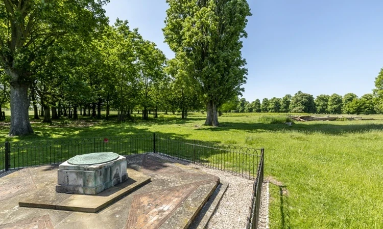 A memorial stone, with a small fence around it, with trees and grass in Bushy Park behind. 