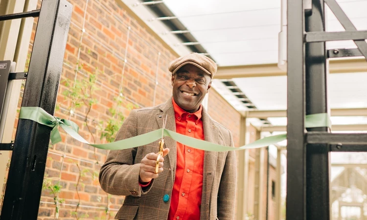 Paterson Joseph is seen looking at the camera whilst cutting a green ribbon across the cafe gate with some gold scissors