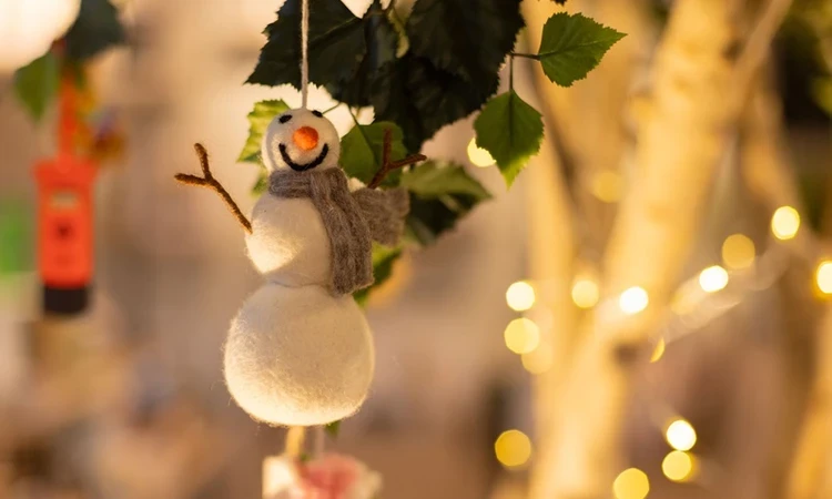 A snowman tree decoration made from felt hangs with fairy lights in the background