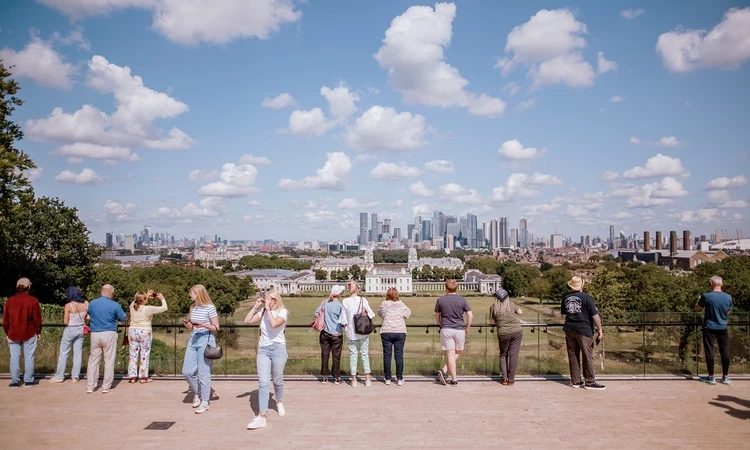 A view from the General Wofle statue looking down toward the Queen's House. White clouds pepper a blue sky and people stand with their back to the camera as they look down at the view