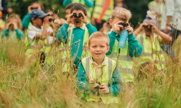  group of young children wearing high-visibility vests and school uniforms are exploring nature in a grassy field. Some of them are using binoculars to observe their surroundings, while one boy in the foreground smiles at the camera, holding binoculars in his hands. The background shows more children and adults, also dressed in high-visibility vests, engaged in the outdoor activity.