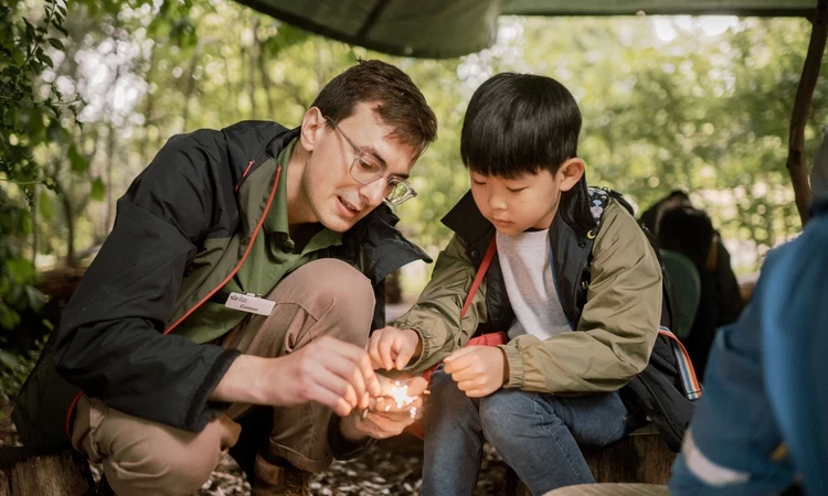 Park ranger and a young child using rocks to generate a fire in an outdoor setting.