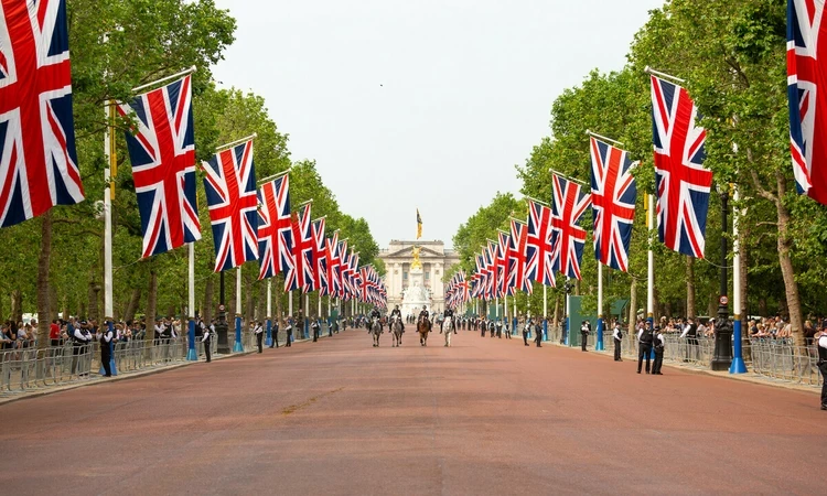 A view of The Mall, looking towards the Queen Victoria Memorial. Both sides of the road are lined with Union Jack flags on flagpoles and trees. 