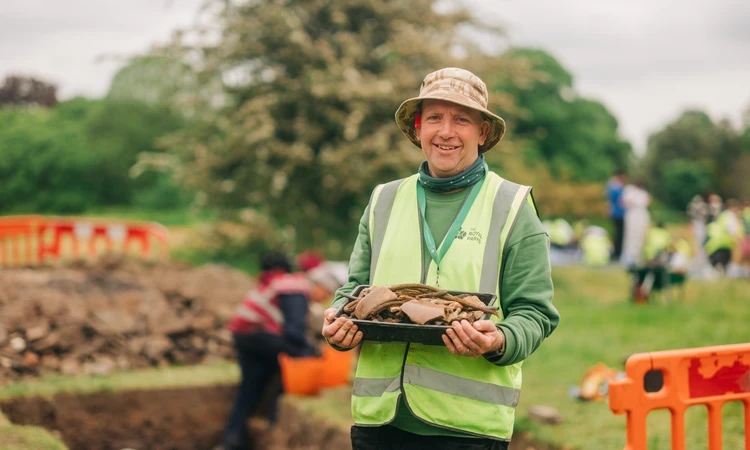 Image of an archaeologist holding onto historical artefacts in an outdoor, park setting with people working and digging in the background.