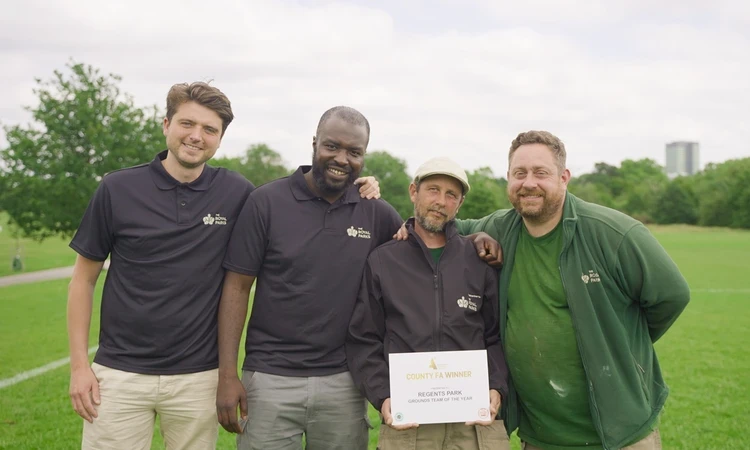 Four men are looking into the camera in front of sports pitches. One of them is holding up a certificate.