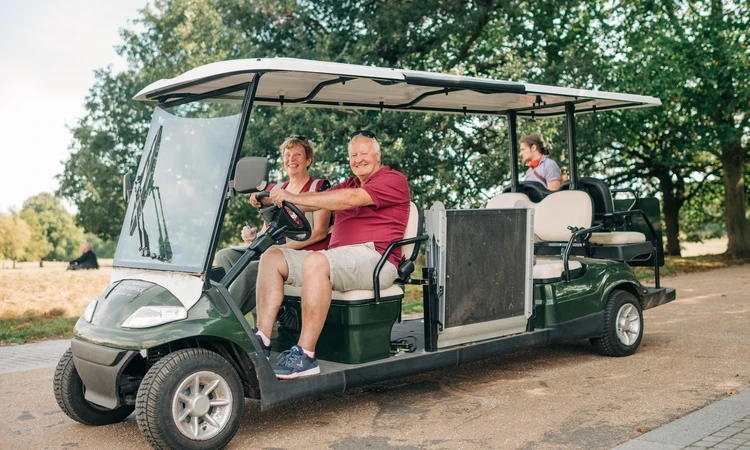 Smiling couple sitting at the front of an open electric buggy in a park, with another passenger seated behind them, surrounded by trees on a sunny day.