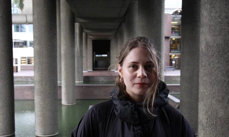Leah, a white woman in her 30s, smiles and looks at the camera. She is wearing a large black coat and stands underneath a bridge or other structure supported by concrete pillars.