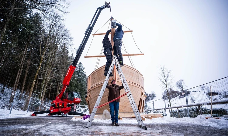 Two men work up a ladder to fit the bowsprit to the galleon, while a third man holds the ladder still. The ground and surrounding trees are snow-covered, and a crane holds the bowsprit in mid-air.