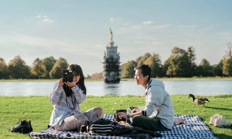 A couple picnicking and taking photography in Bushy Park, with the Diana fountain in the background