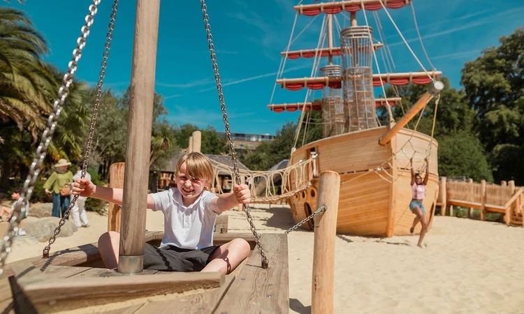 A boy sits in the wooden play boat with the new galleon behind