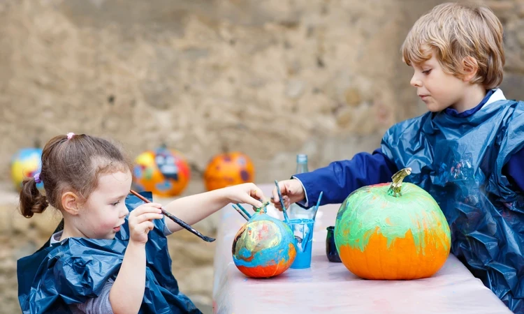 Two children sit either side of a table. They are wearing blue plastic aprons and have paintbrushes in their hands directed at large orange pumpkins that are being colourfully painted. 