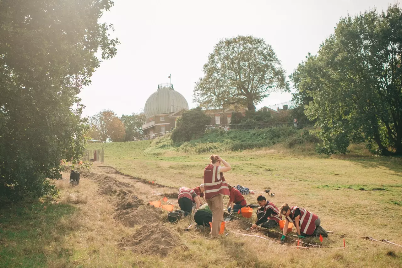 A wide angle shot of the trench being dug by volunteers looking uphill with the Royal Observatory in the background. 