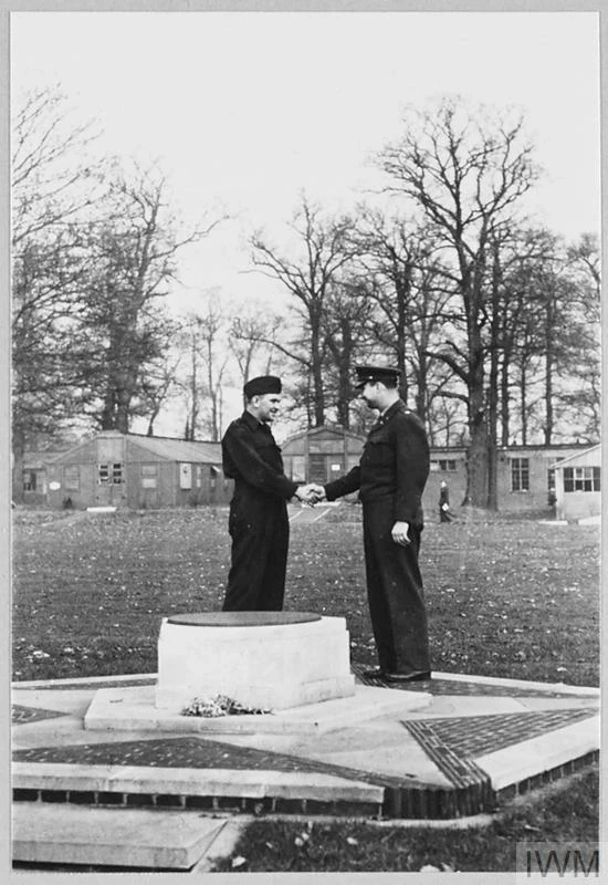 Two people shaking hands in front of the USAAF memorial. Huts can be seen alongside trees in the background.