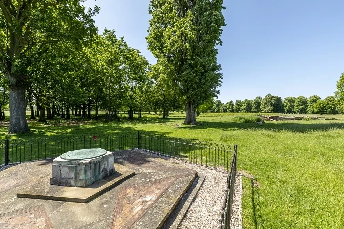 A memorial stone, with a small fence around it, with trees and grass in Bushy Park behind.
