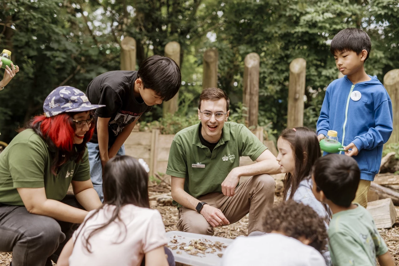 A male teacher/ park ranger talking to a group of children sitting around him in a circle. They are in an outdoor setting.