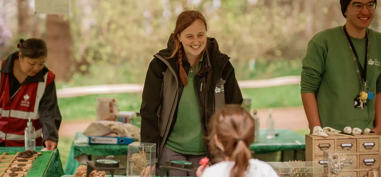 Image of 3 adults behind a desk, in an outdoor setting, explaining educational activities to a child.