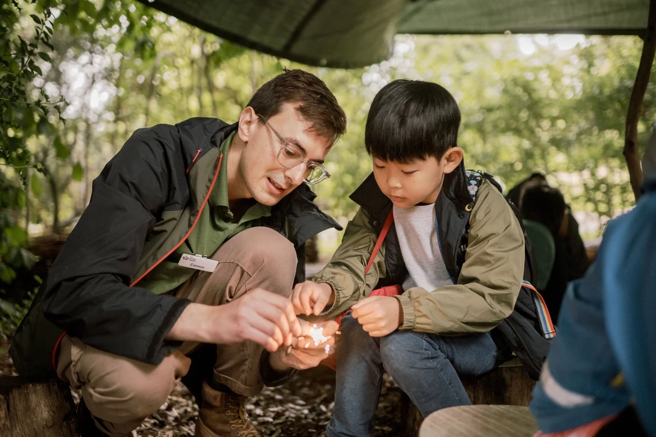 Park ranger and a young child using rocks to generate a fire in an outdoor setting.