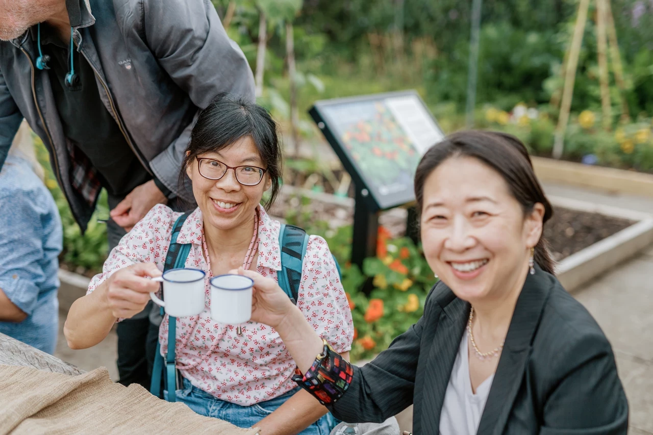 Two people sitting in an allotment smile at the camera as they lift their cups of herbal tea in the air in celebratory style