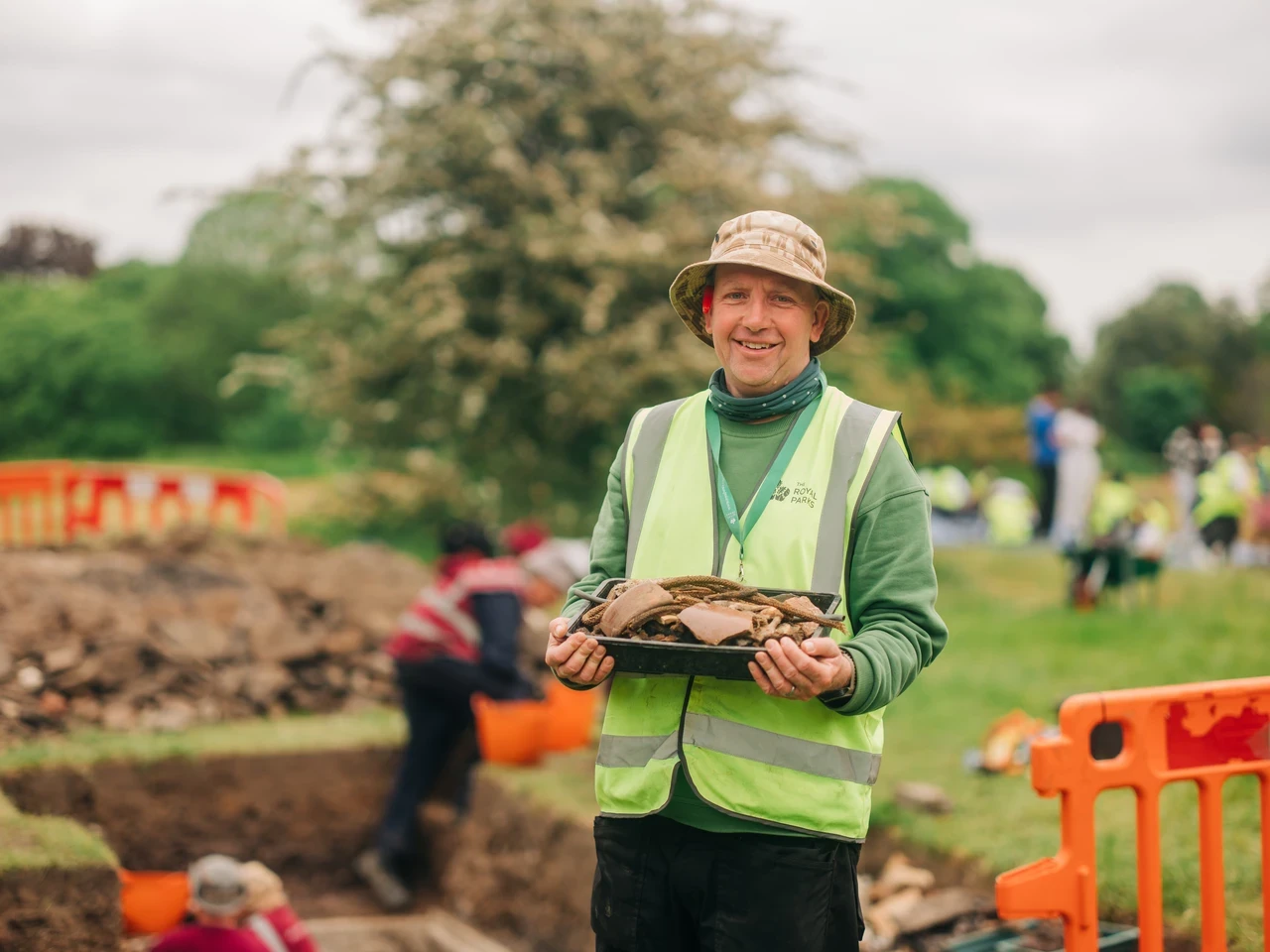 Image of an archaeologist holding onto historical artefacts in an outdoor, park setting with people working and digging in the background.
