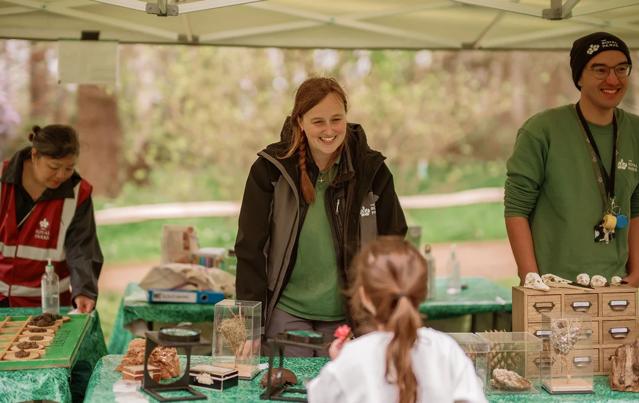Charlotte Cass pictured outdoors in a Royal Parks Jumper and black coat, across from a table of natural learning resources from a young learner