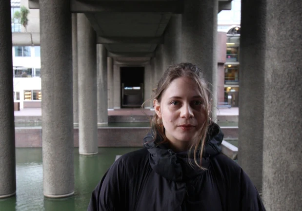  Leah, a white woman in her 30s, smiles and looks at the camera. Her dark blonde hair is tied up but loose and windswept. She is wearing a large black coat and gold earrings, behind her are rows of concrete pillars receding into the background, holding up a roof over a green body of water.