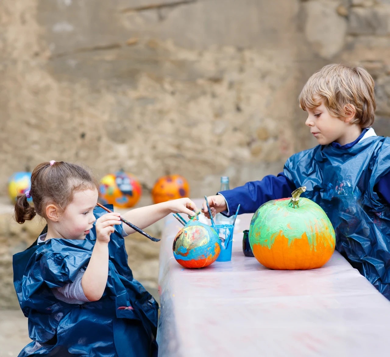 Two children sit either side of a table. They are wearing blue plastic aprons and have paintbrushes in their hands directed at large orange pumpkins that are being colourfully painted. 