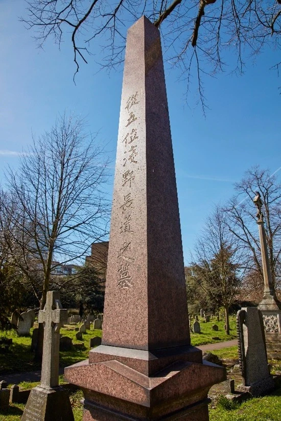 Tall obelisk style brown grave amidst a green graveyard.